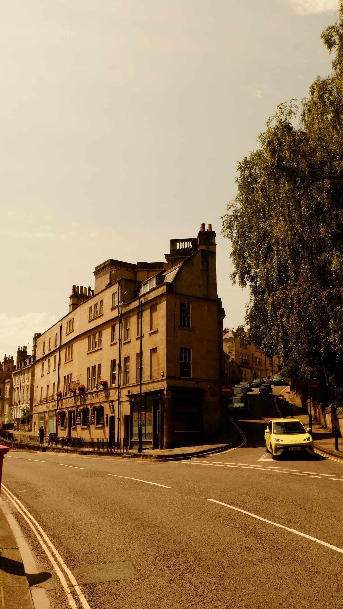 A yellow car coming down a curving hill road past a tall stone corner building and a large tree