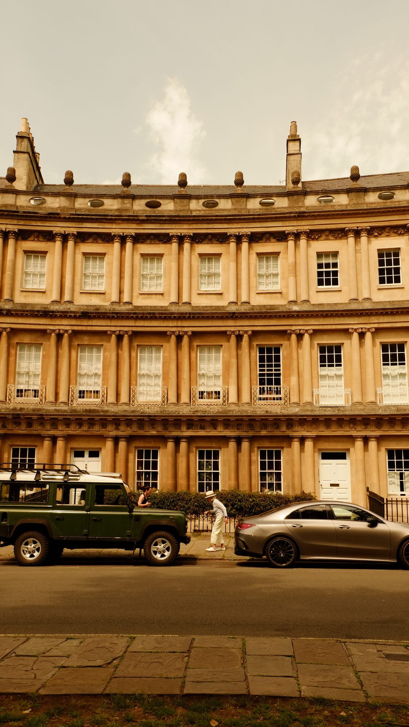 A green Land Rover and a grey coupé parked below the columned façade of the Circus as a man in a white hat walks by