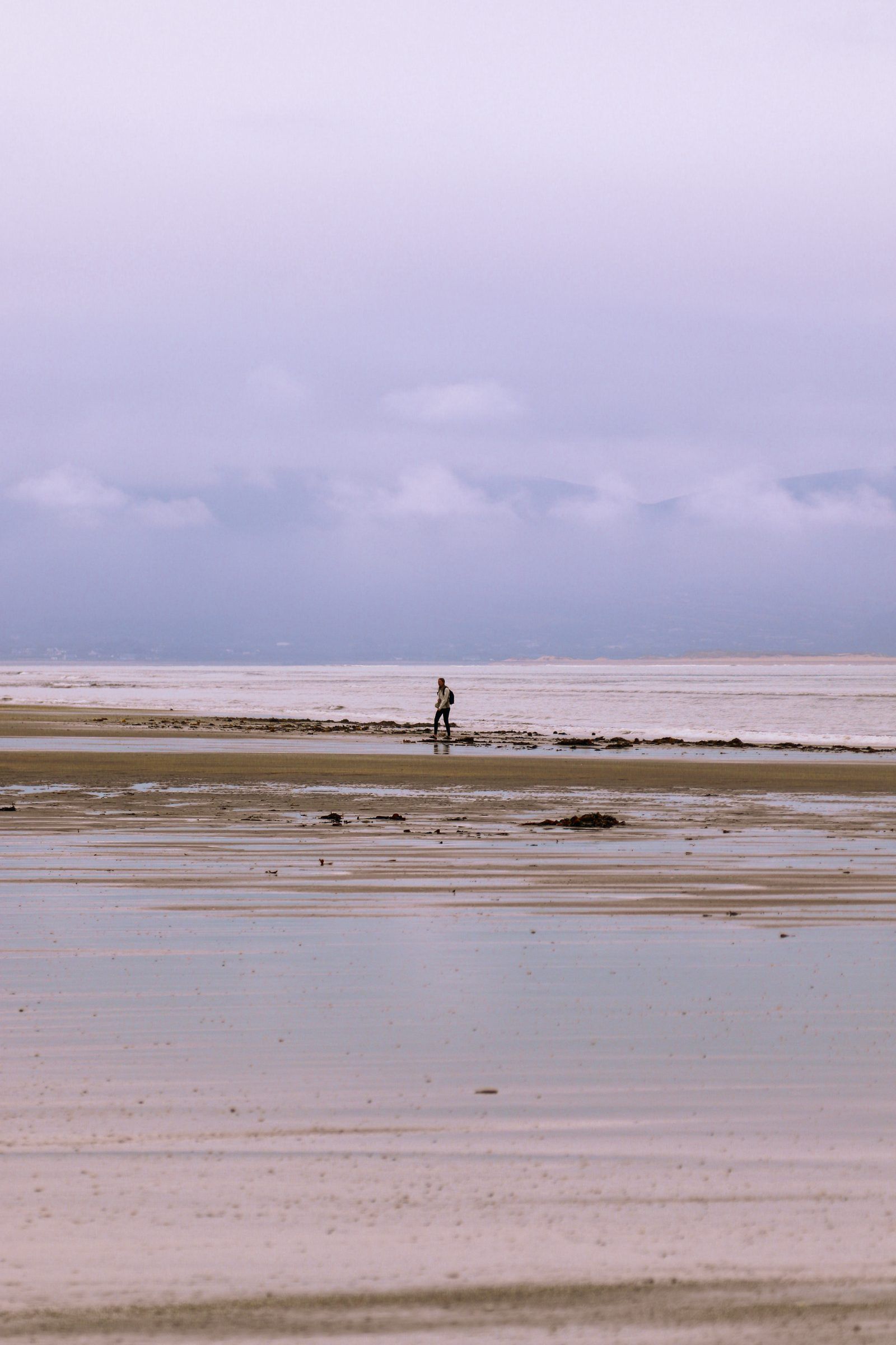 A lone figure standing far out on a vast wet beach under a pale lilac sky