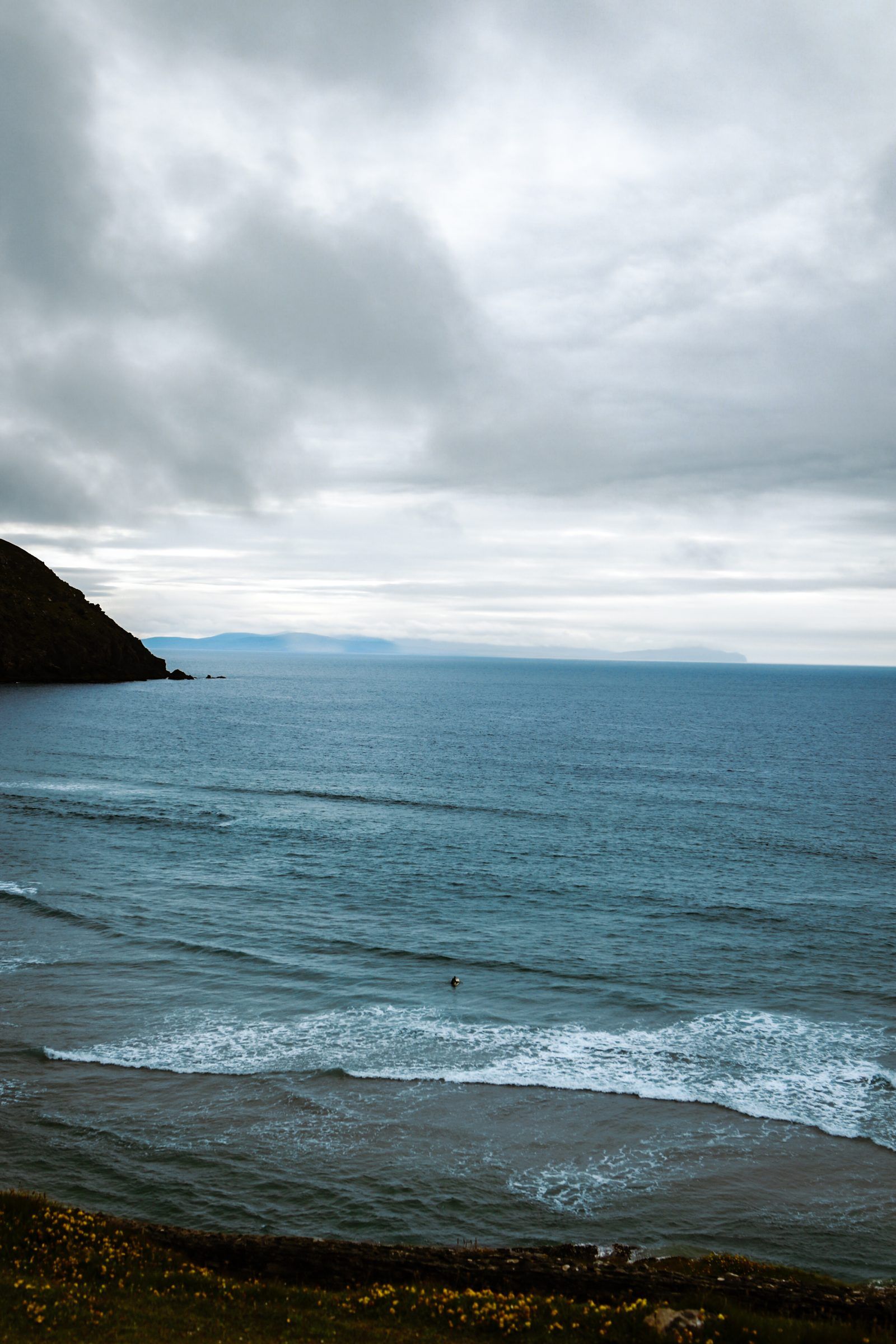 A single surfer far out in a dark blue sea as a line of white surf rolls in, a headland under heavy cloud