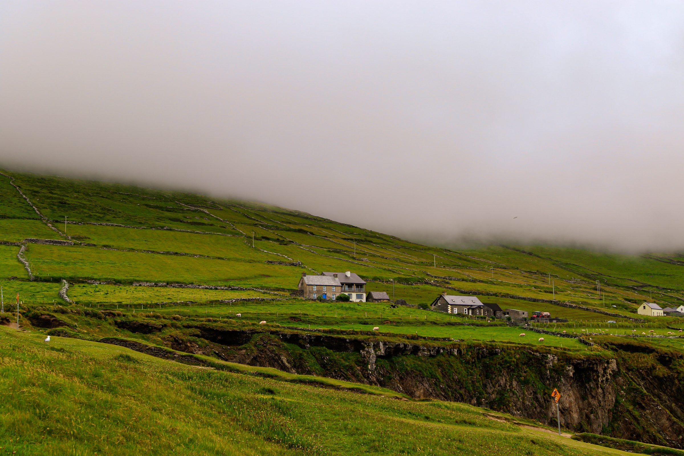 Stone cottages on a green hillside above a cliff edge, low cloud hiding the top of the slope