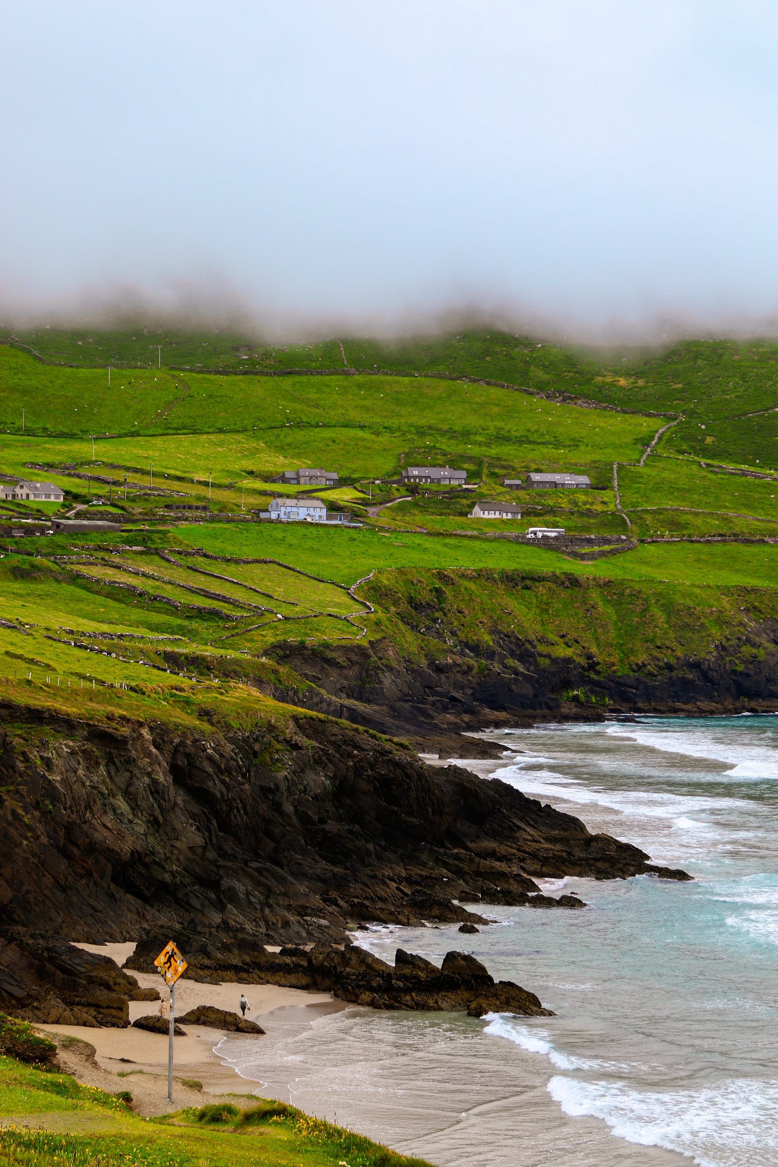 Waves rolling into a cove below dark cliffs, a road sign on the beach path and farms on the green slopes above