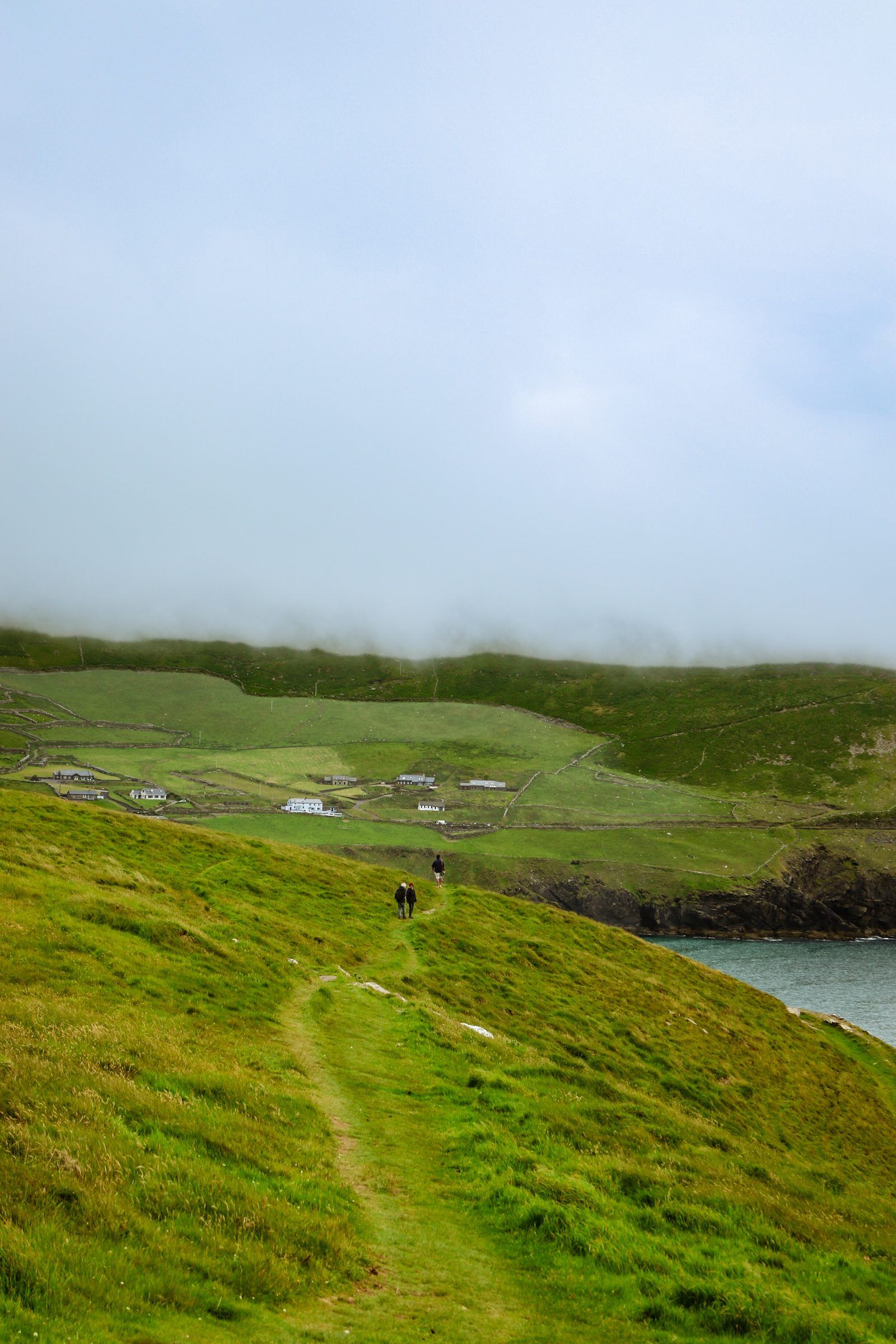 Two walkers on a grassy path leading down to white farm buildings, fog lying low over the hills