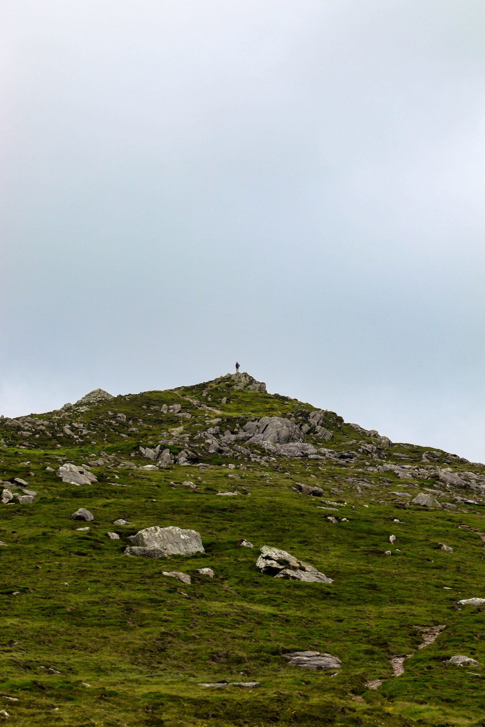 A tiny figure standing on top of a rocky, grass-covered hill beneath a pale overcast sky