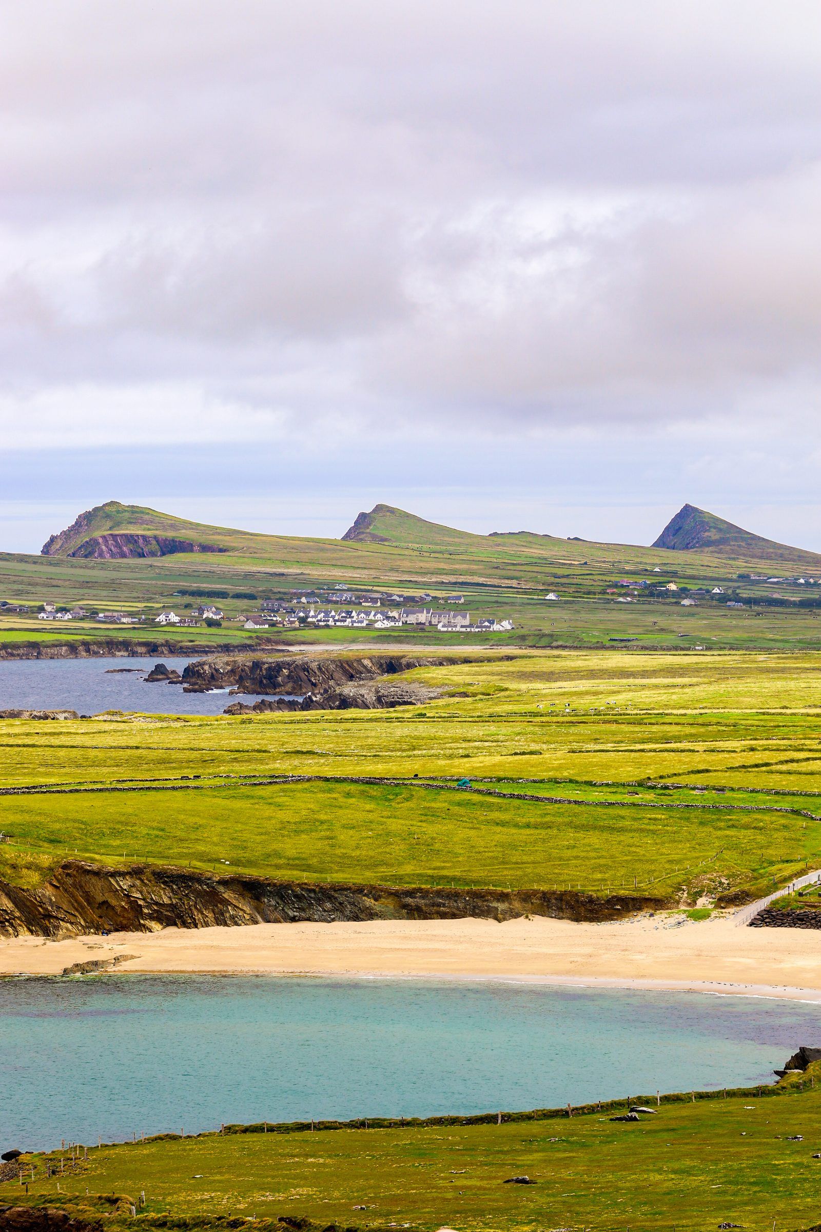 A sandy cove with turquoise water below green fields, three pointed headlands rising along the horizon
