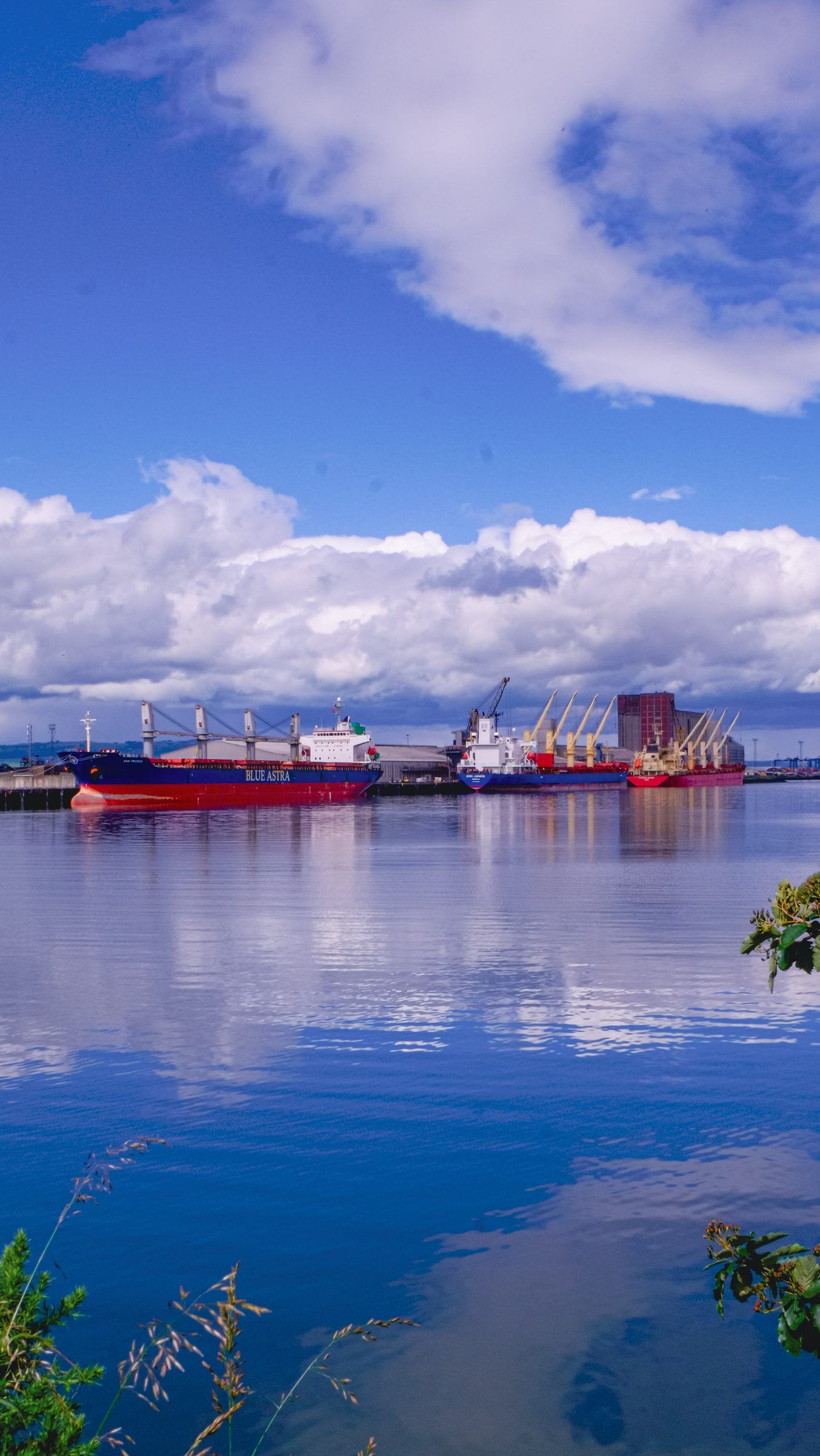 Cargo ships and cranes reflected in vivid blue harbour water under bright white clouds