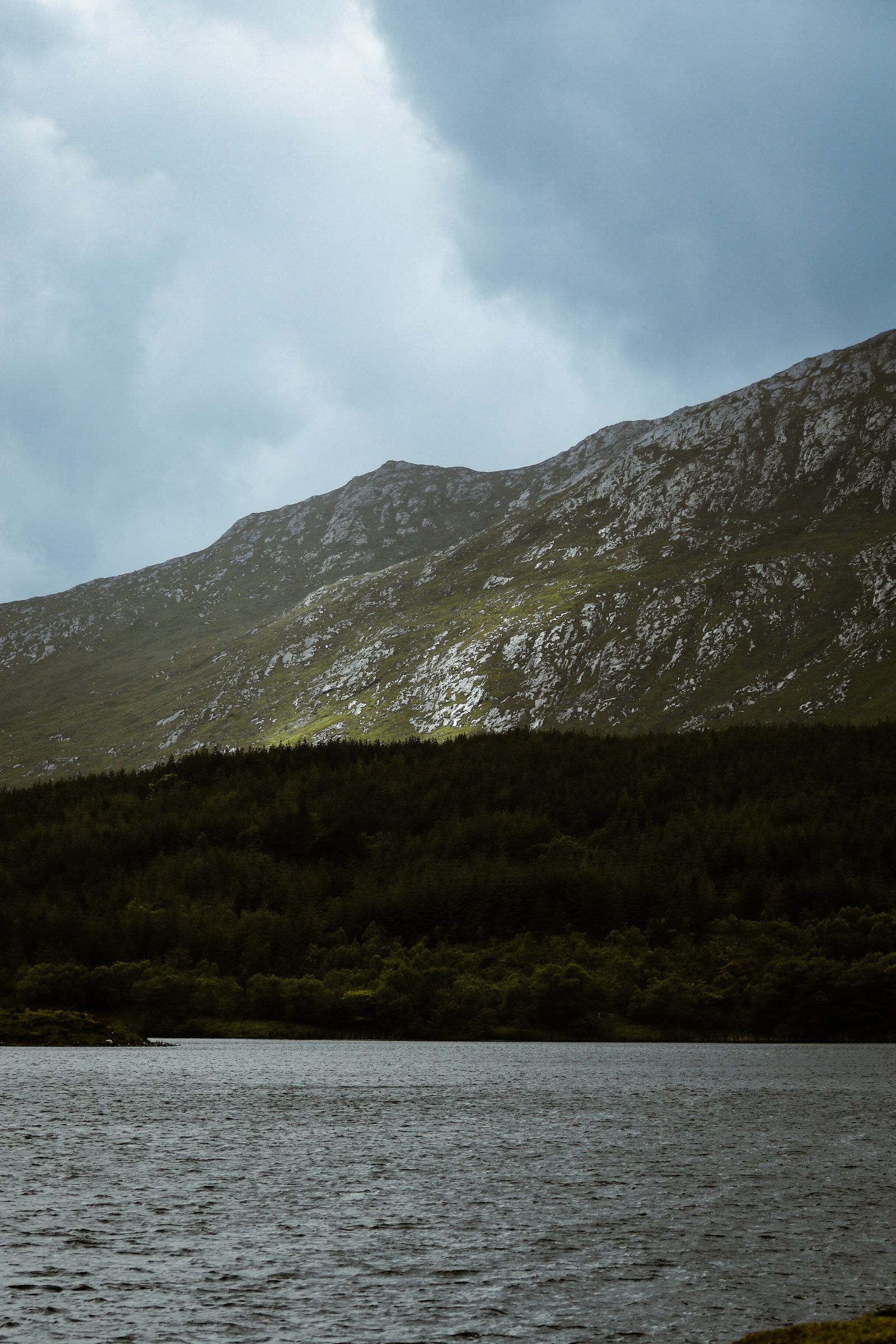 A rocky grey mountain rising above a dark band of forest and a wind-ruffled lake under storm clouds