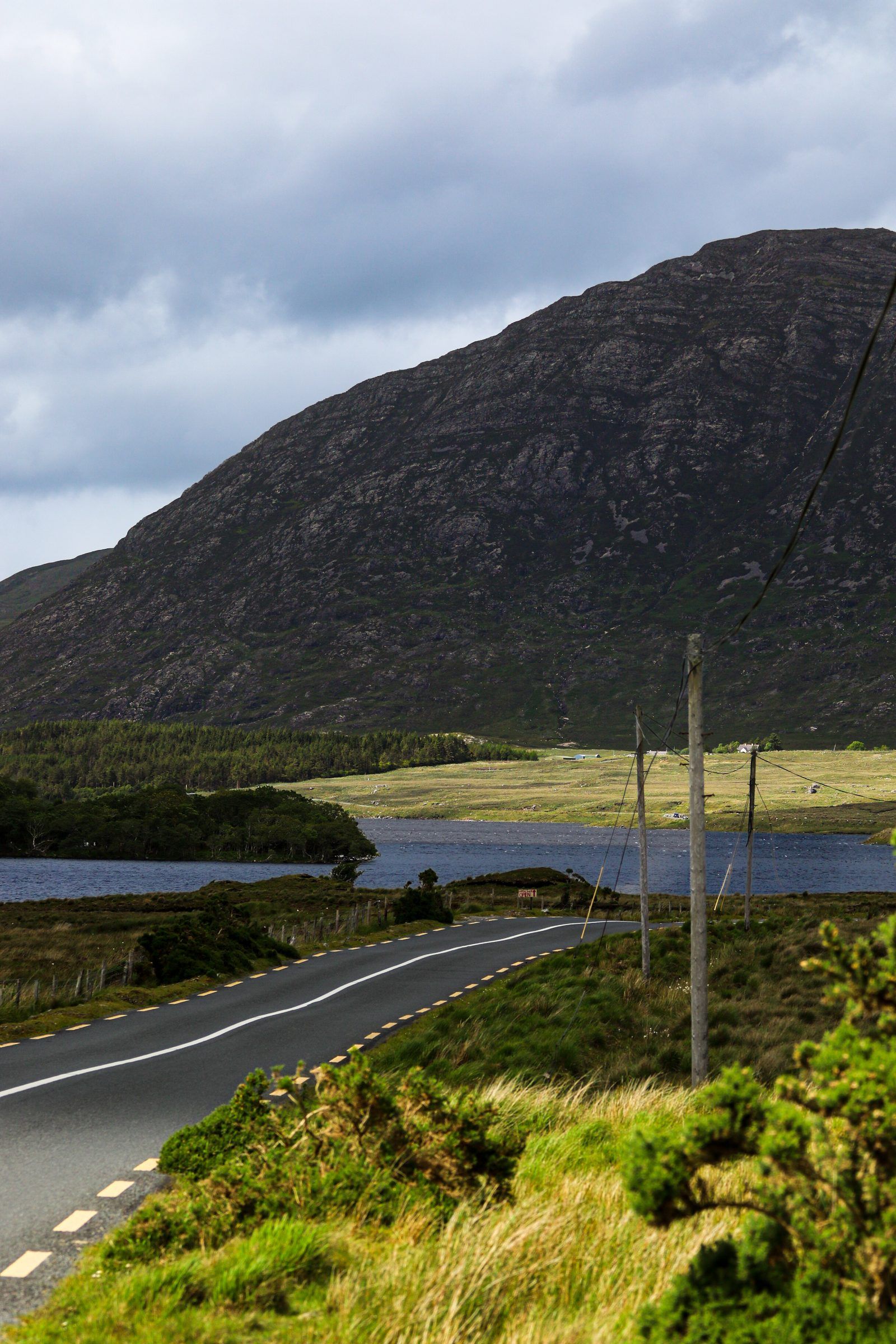 An empty road curving past telegraph poles toward a lake and a dark, bare mountain