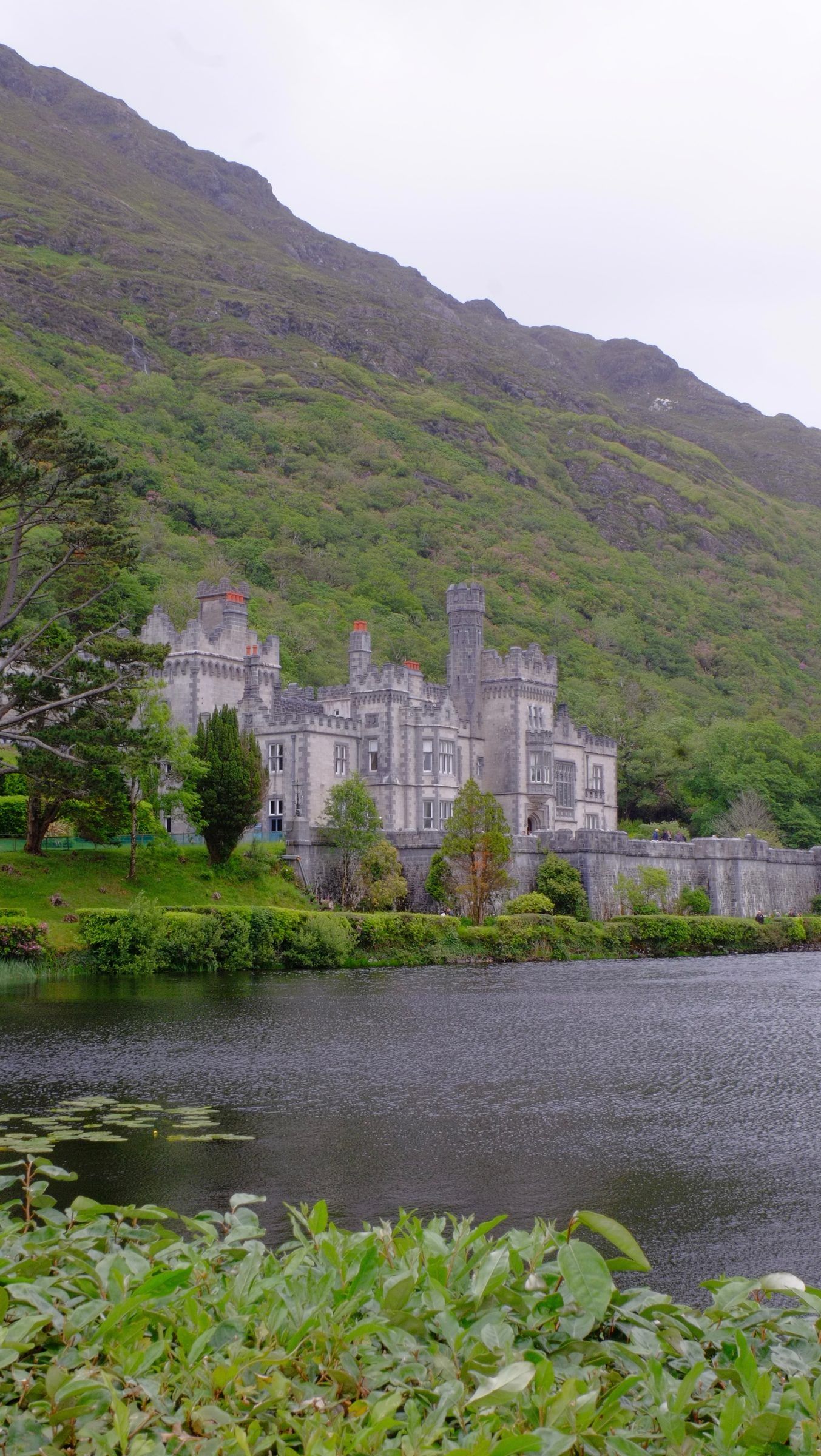 A grey castellated abbey at the foot of a steep green mountainside, seen across a dark lake fringed with leaves