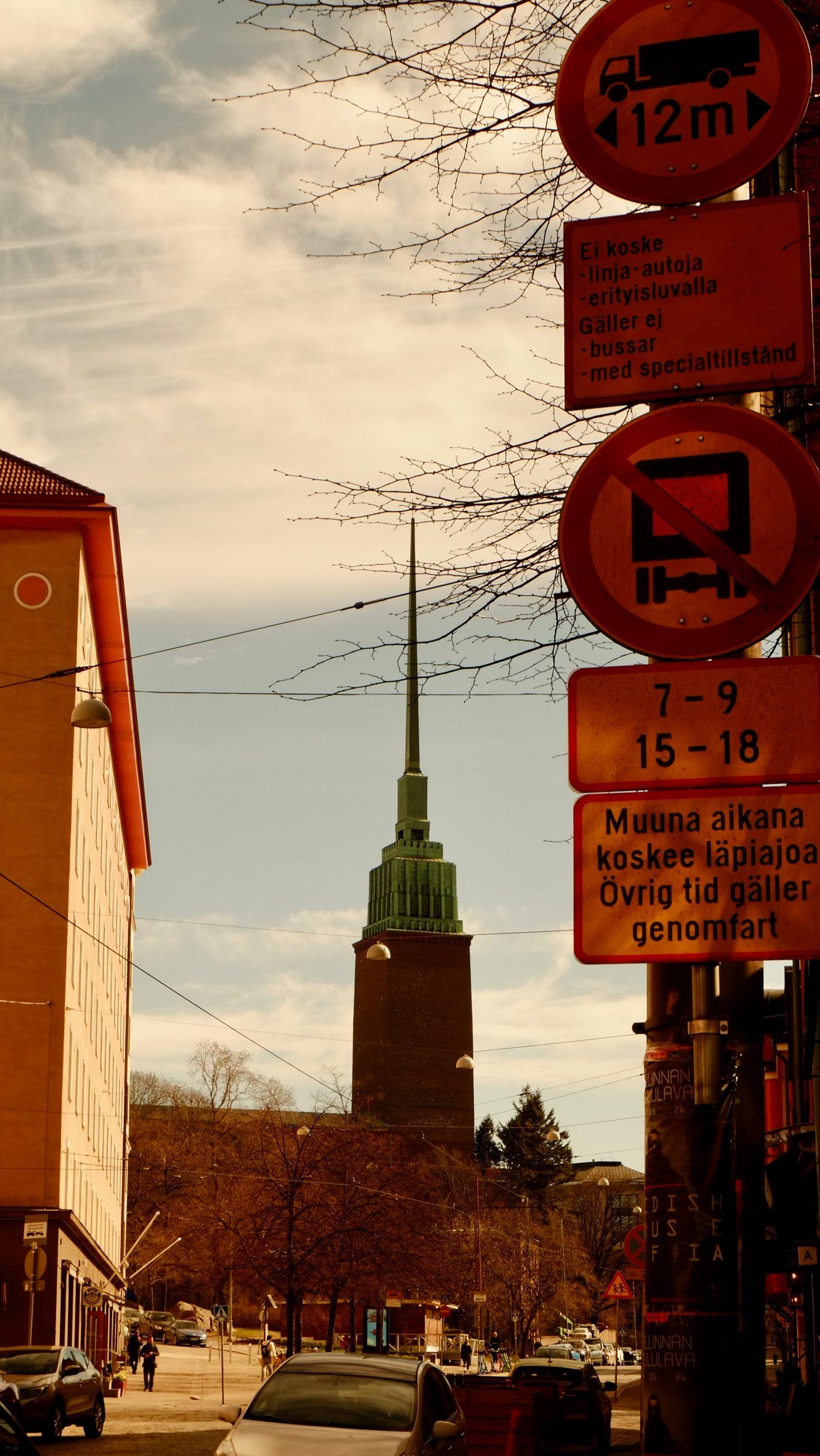 A stack of red and yellow traffic signs beside a street leading to a brick church tower with a slender green spire