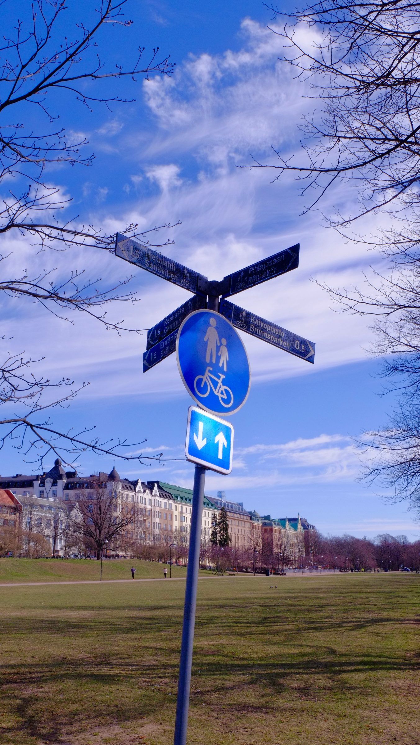 A blue cycle and footpath sign beneath street-name blades in a park, a row of grand apartment buildings beyond