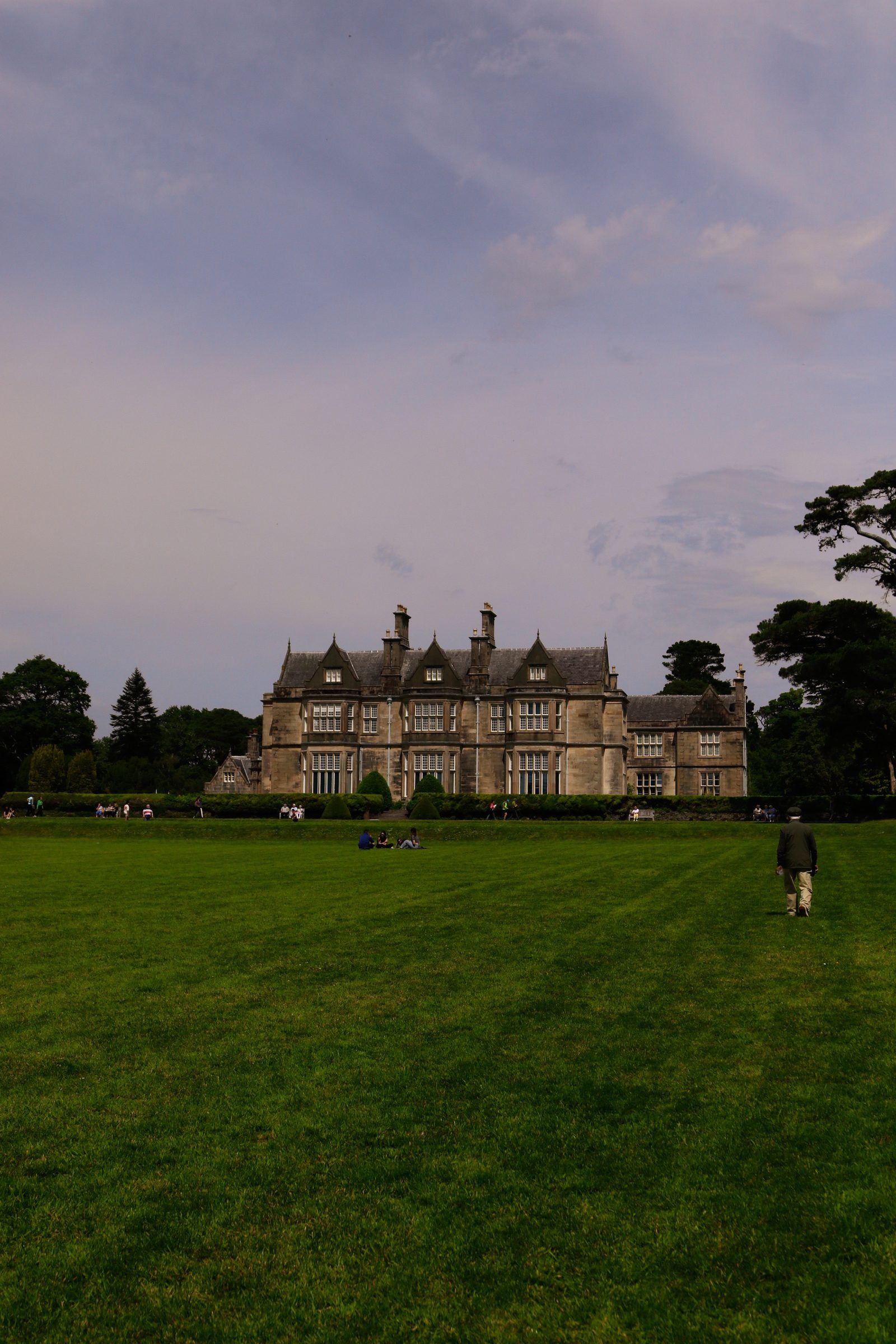A lone walker crossing a broad striped lawn toward a distant gabled mansion under a heavy sky