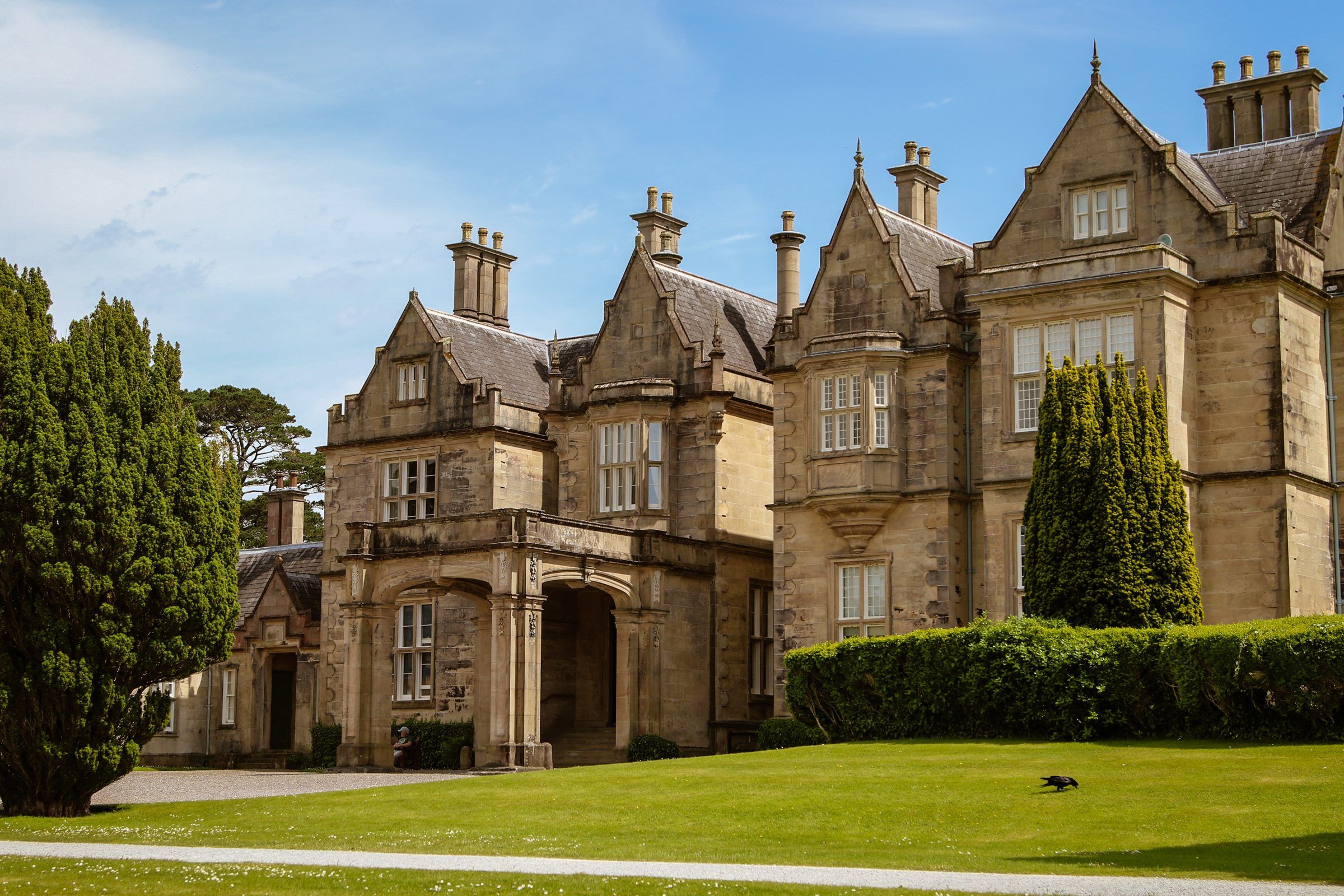 A sandstone Victorian mansion with tall gables and chimneys beside clipped hedges and dark yews under a blue sky