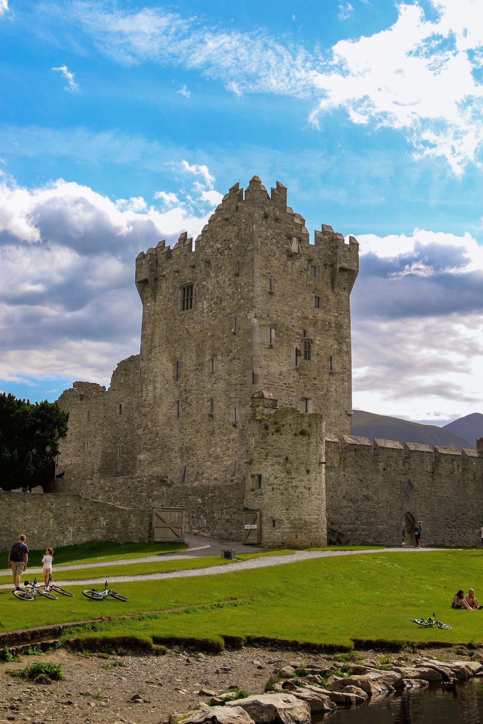 A stone tower house castle with battlements beside a lake shore, people and bicycles resting on the grass