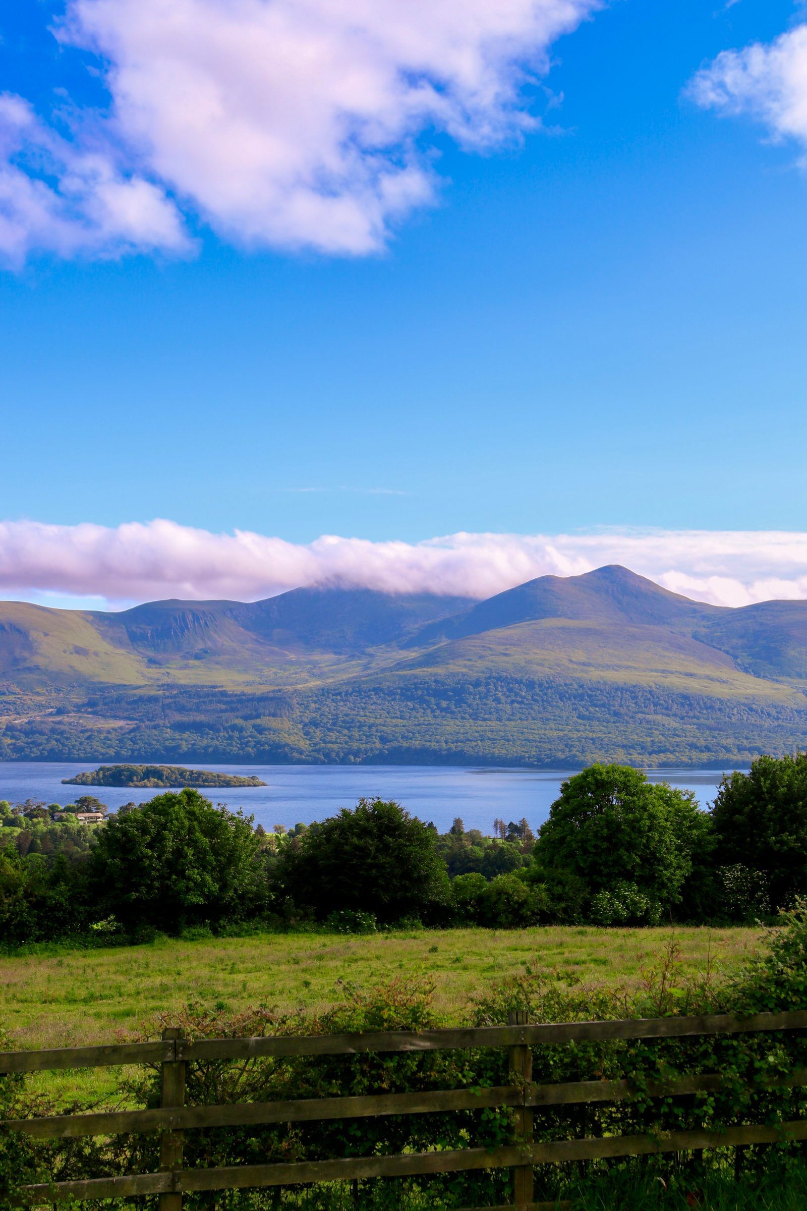 A blue lake with a wooded island beneath green mountains and a band of white cloud, a wooden fence in the foreground
