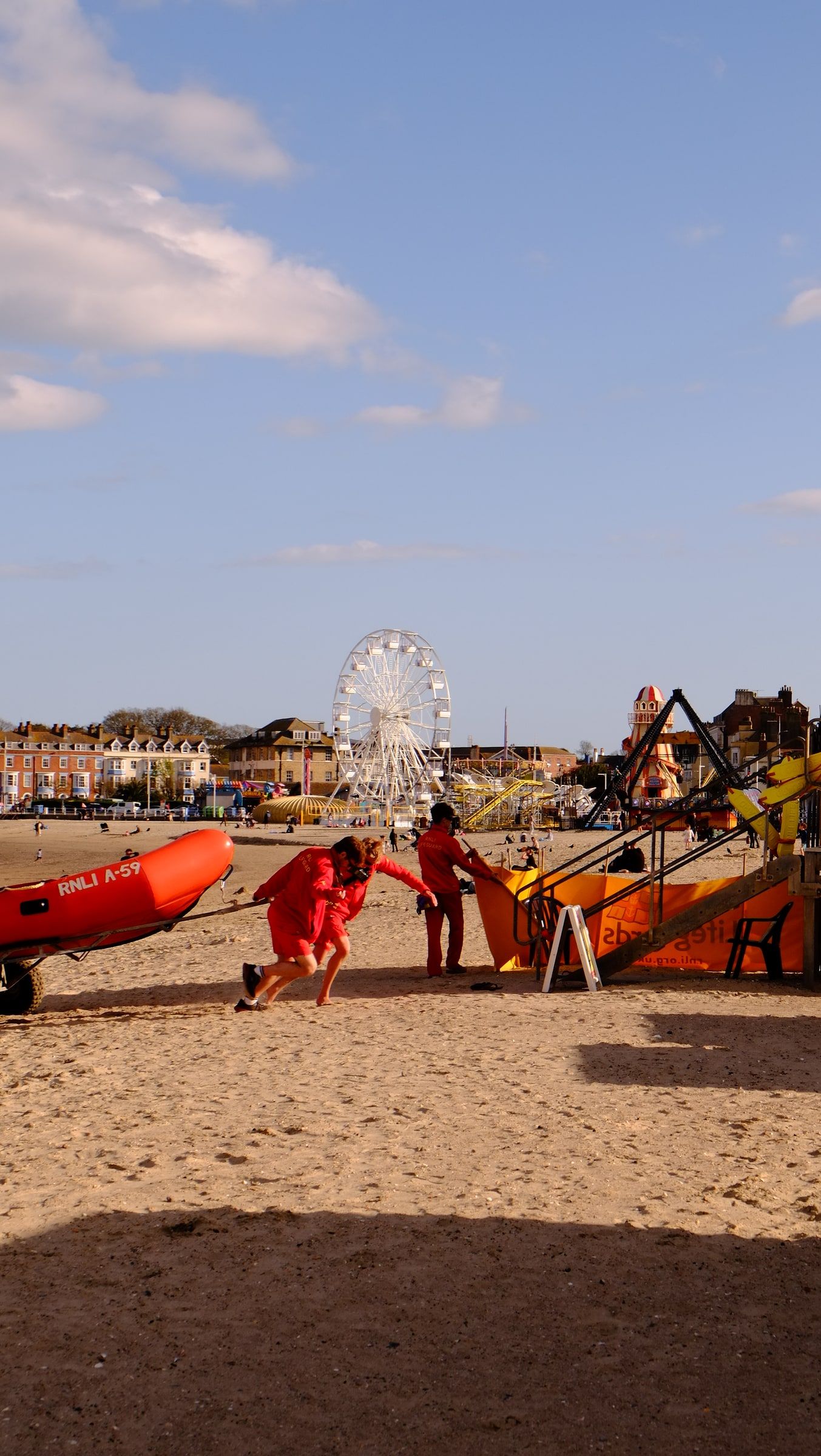 Lifeguards in red hauling an inflatable rescue boat across Weymouth beach, a Ferris wheel and the seafront behind