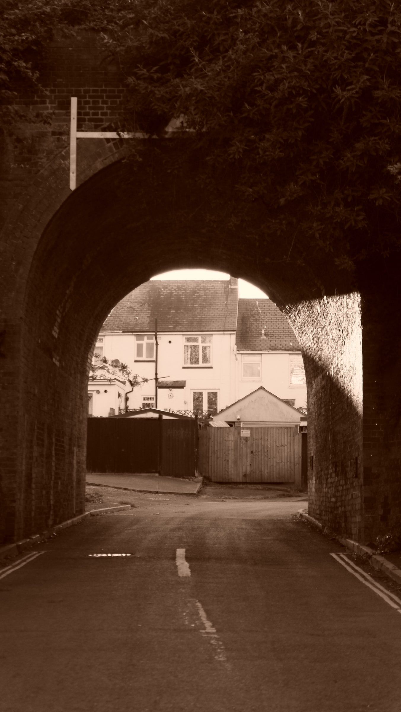 A sepia view through a dark railway arch to a white cottage and a quiet lane beyond