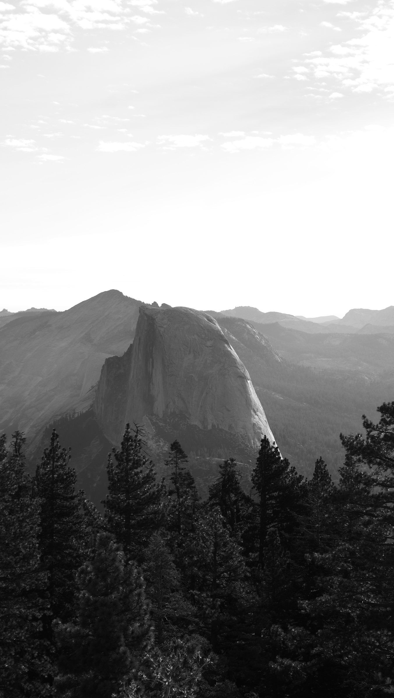 A black-and-white view of Half Dome rising above dark pines under a bright hazy sky