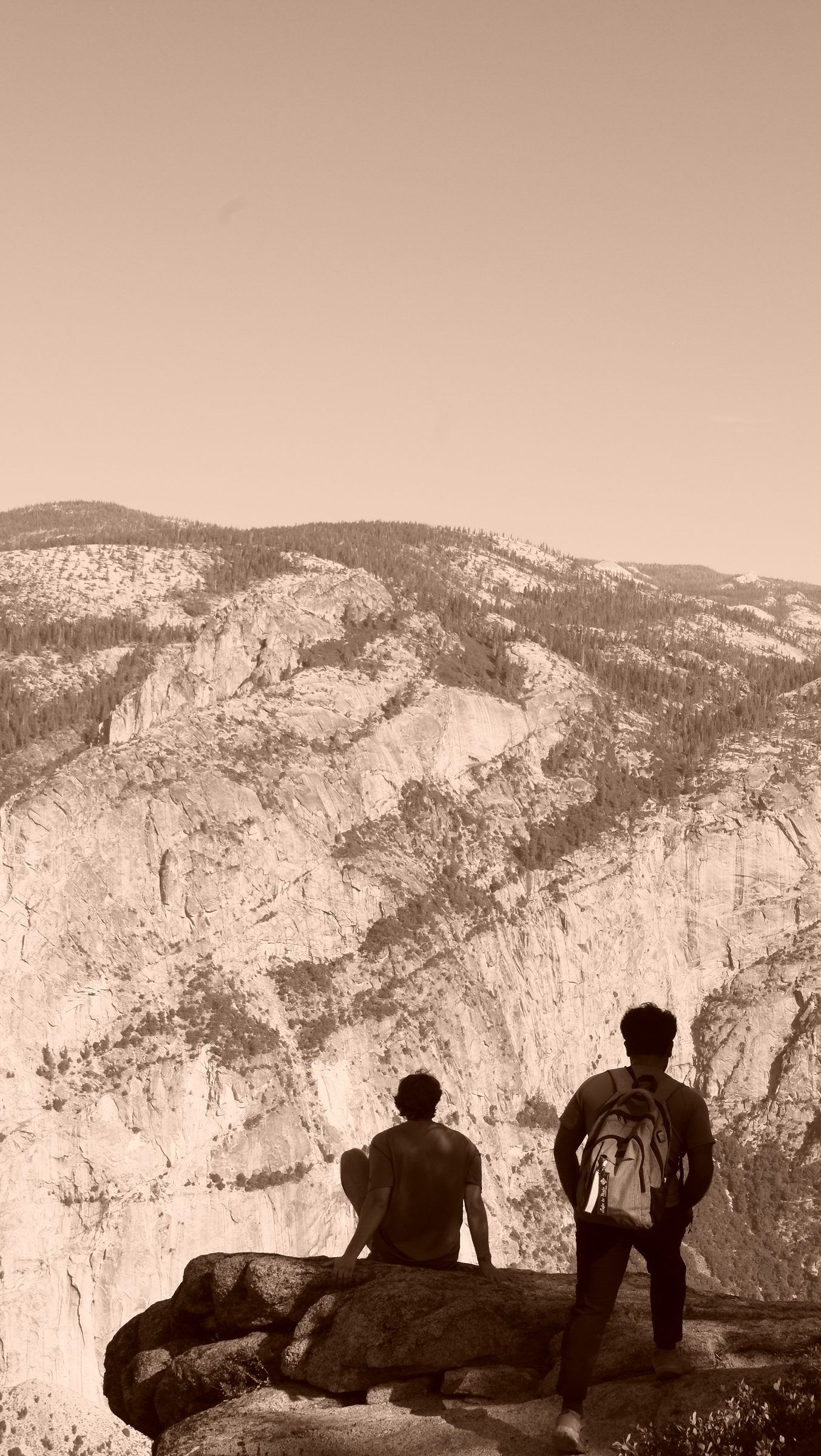 A sepia view of two hikers, one seated and one standing, on a rock ledge facing a vast granite wall