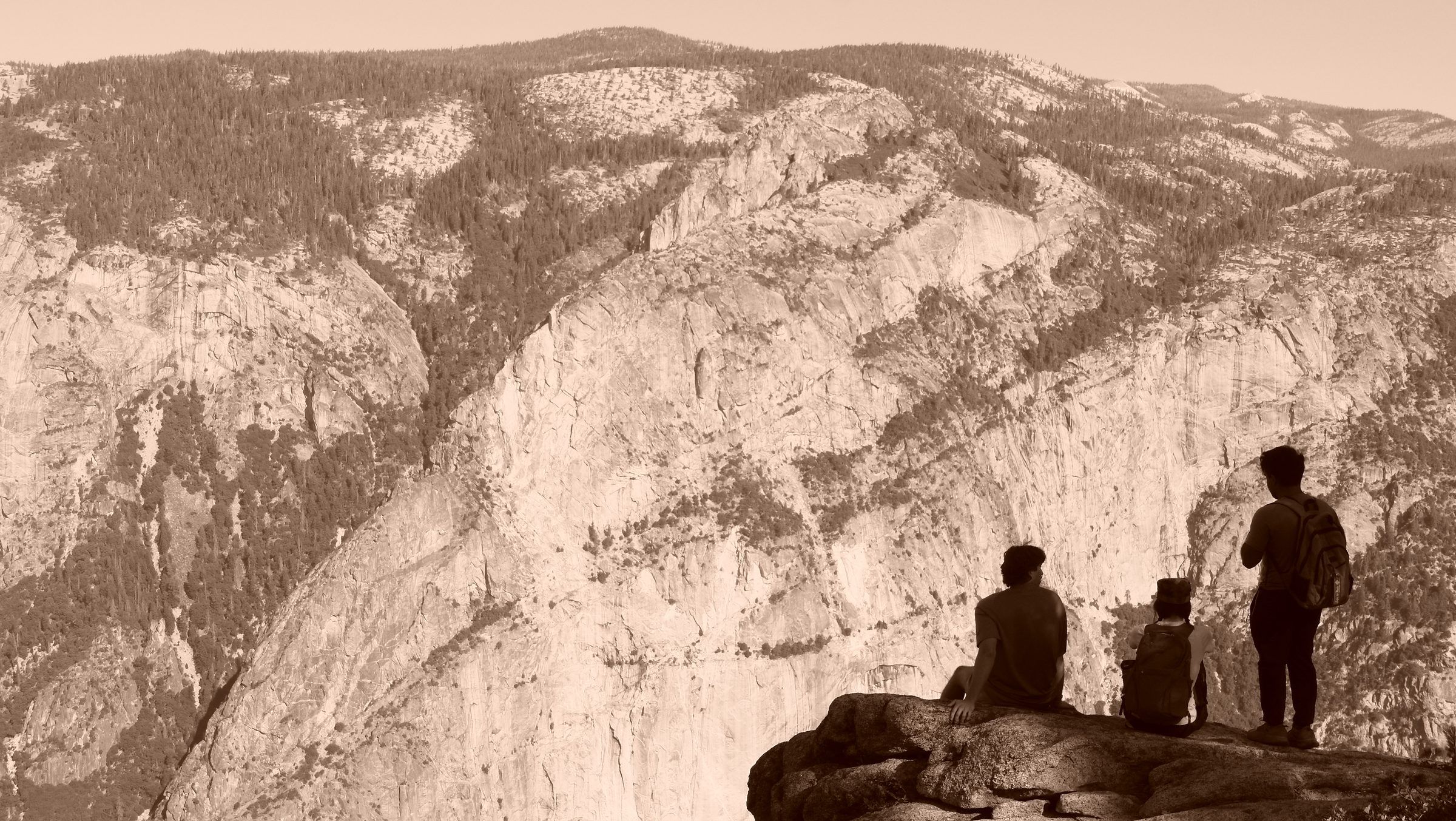 A sepia view of three hikers resting on a rocky outcrop, dwarfed by a granite mountainside dotted with trees